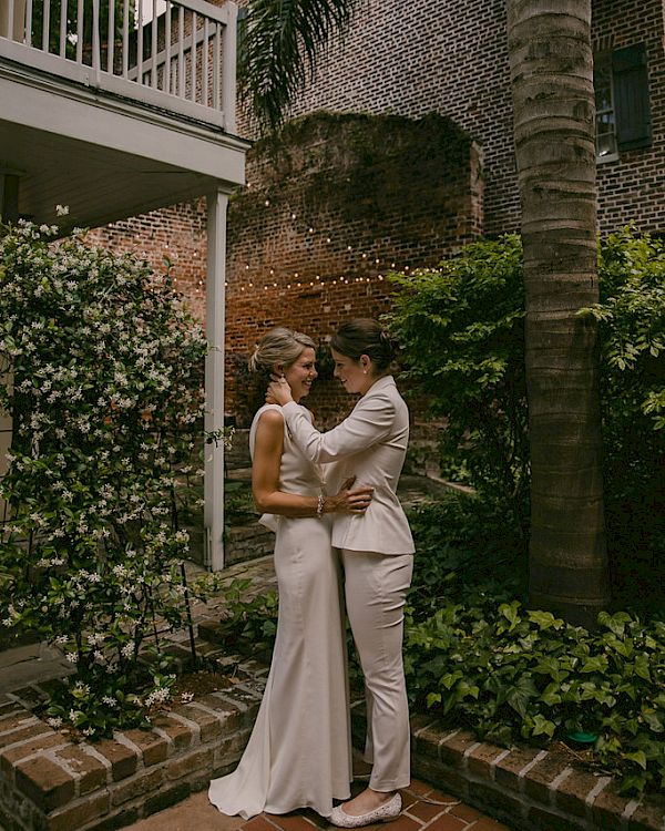 Two people in wedding attire lovingly gaze at each other in a lush garden setting with a brick wall and string lights in the background.