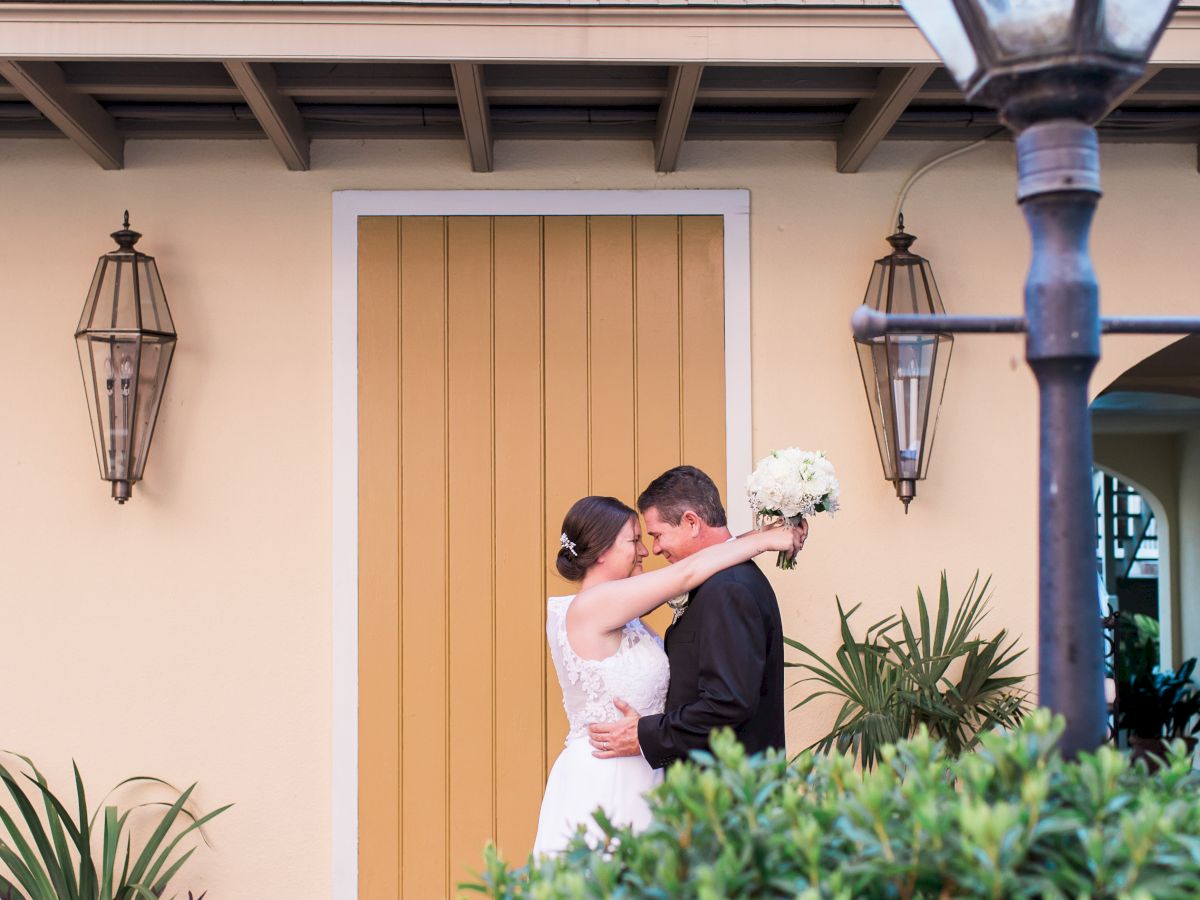 A couple embraces in front of a yellow door, with a bouquet, surrounded by greenery and lanterns under a balcony.
