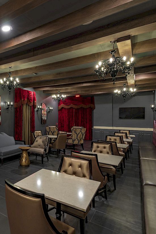 A stylish room features elegant chandeliers, ornate curtains, artwork, cushioned chairs, and tables, creating an opulent ambiance.