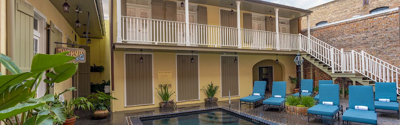 A courtyard with a pool, patio chairs, potted plants, and a balcony in a building with two floors and brick accents.