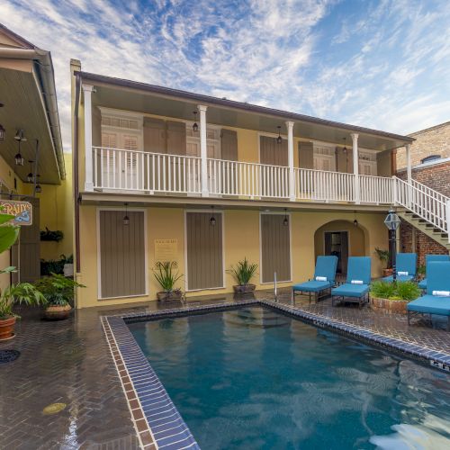 A courtyard with a pool, patio chairs, potted plants, and a balcony in a building with two floors and brick accents.
