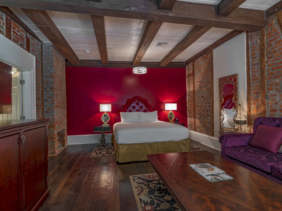 A stylish bedroom features a red accent wall, cozy bed, velvet couch, wooden beams, and elegant decor for a luxurious atmosphere.