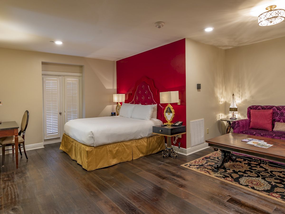 A luxurious bedroom with a bed, ornate headboard, vibrant red accent wall, purple sofa, wooden table, and elegant decor.