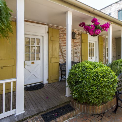 A charming porch with brick walls, yellow shutters, and hanging plants, featuring a bench and two doors labeled "109", surrounded by shrubs.