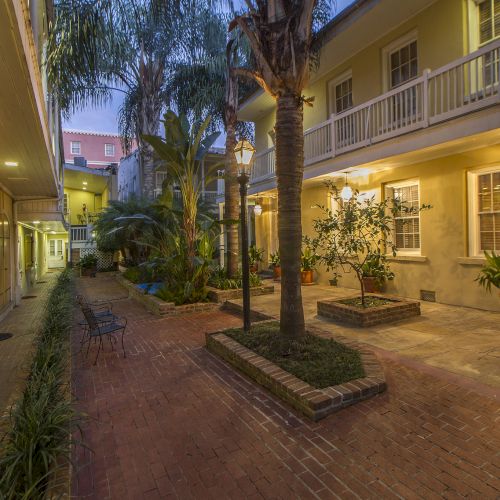 A cozy courtyard with brick pathways, palm trees, plants, and warmly lit buildings. It includes chairs and a lamp post.