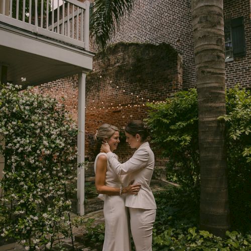 Two people in wedding attire are embracing each other in a garden setting with brick walls and lush greenery.