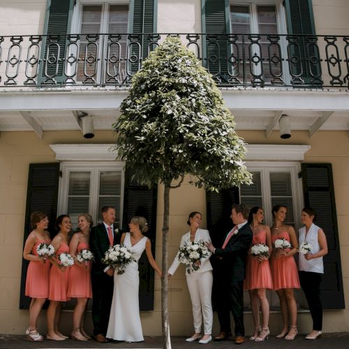 A group of people in formal attire with bouquets, likely a wedding party, stands outside a building with a balcony and a tree in the middle.