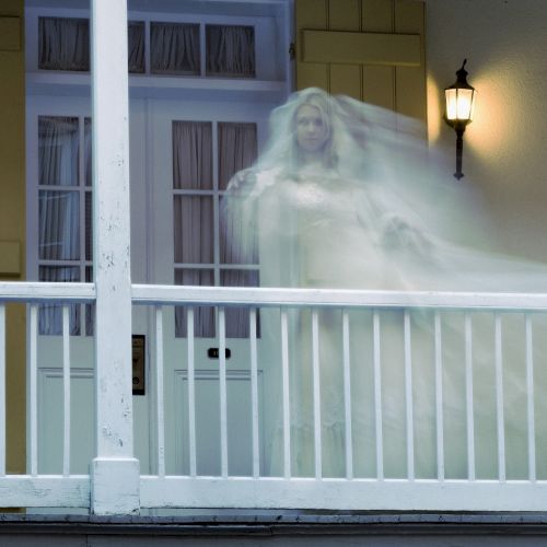 A ghostly figure in white appears on a porch with closed shutters, softly illuminated by a wall-mounted light.
