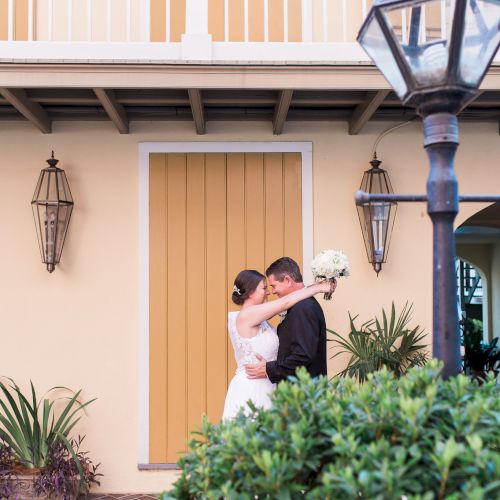 A couple embraces in front of a yellow building with plants and vintage lamps around them.