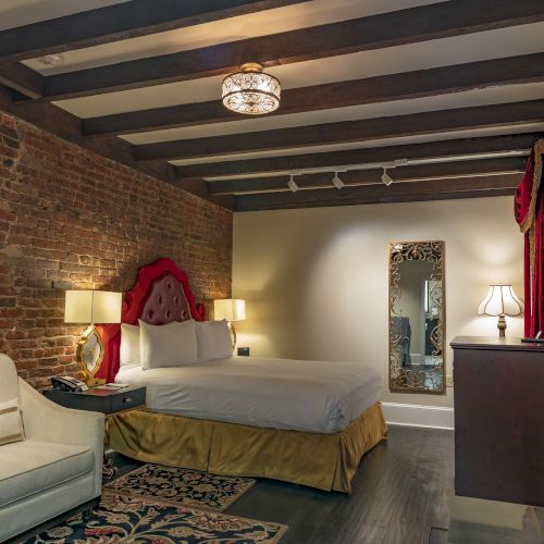 A cozy bedroom with a brick wall, bed with a red headboard, sofa, mirror, and lamps, featuring vintage decor and warm lighting.