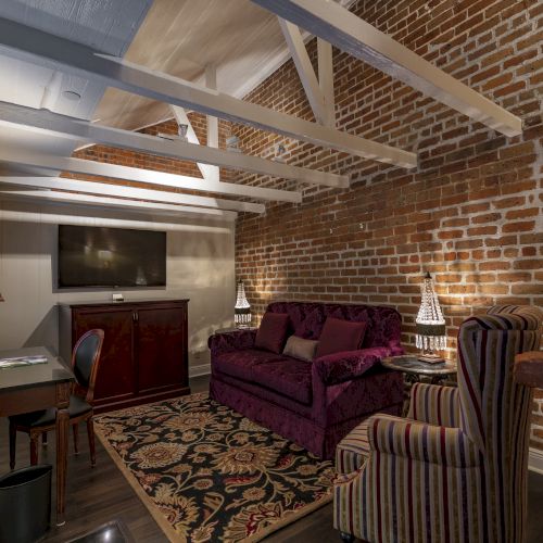 A cozy room with exposed brick walls, a purple sofa, striped chair, TV, desk, and ornate lamps, creating an inviting atmosphere.