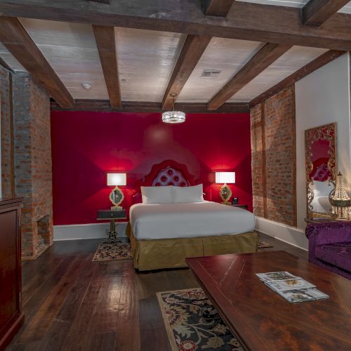 A stylish bedroom with a red accent wall, elegant bed, purple sofa, and exposed brick, creating a blend of rustic and modern design.