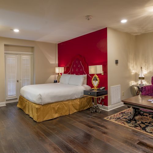 A cozy room with a bed, desk, purple sofa, and wooden floor. Red accent wall, ornate lamps, and a patterned rug add elegance.