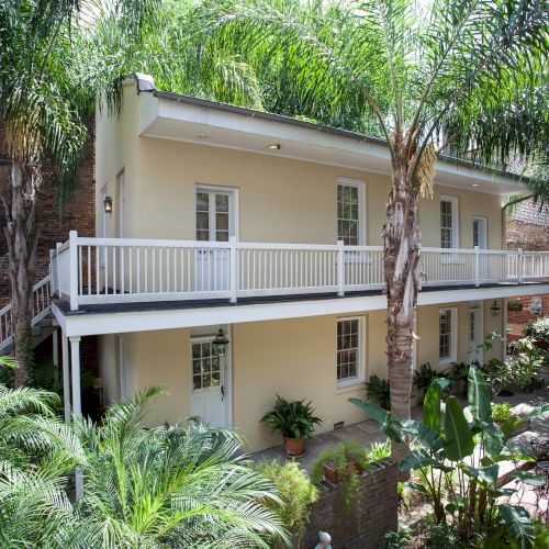 A two-story house surrounded by palm trees, with a brick patio and metal outdoor furniture in the foreground.