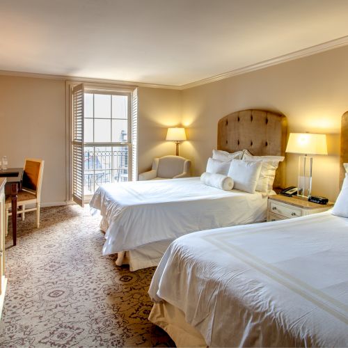 The image shows a hotel room with two large beds, a TV, a desk with chairs, lamps, and an open window with shutters.