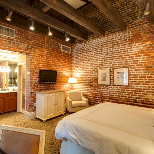 A cozy bedroom with brick walls, a bed, armchair, TV, and a small bathroom area with a mirror and towels.
