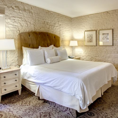 A cozy bedroom with a brick wall, white linens, upholstered headboard, side tables, lamps, artwork, and a patterned carpet.