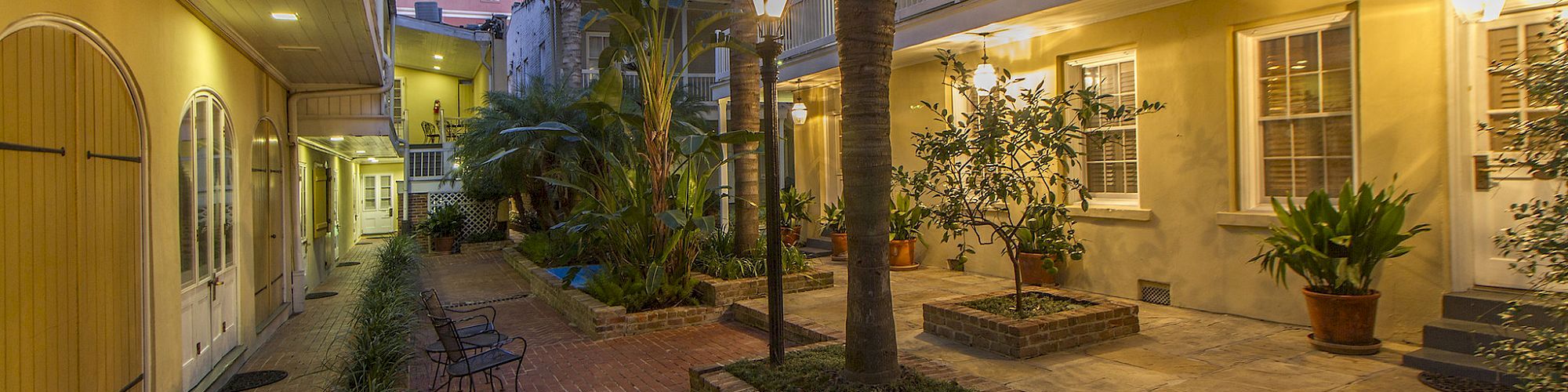 A charming courtyard with brick paths, plants, and trees, surrounded by two-story yellow buildings with balconies and warm lighting.