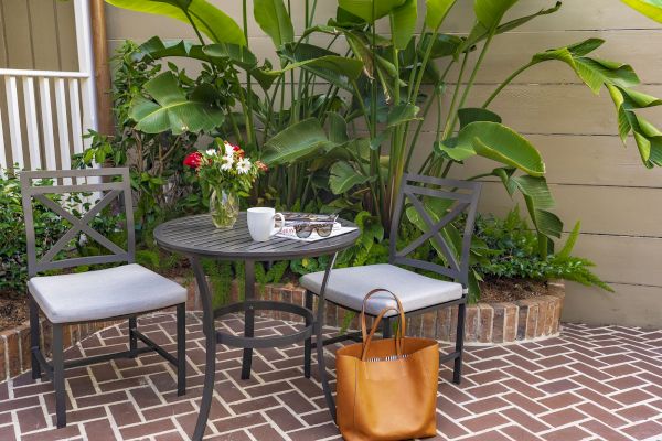 A patio scene with two chairs, a round table holding a mug, sunglasses, and a flower vase; surrounded by lush green plants and a brown bag.