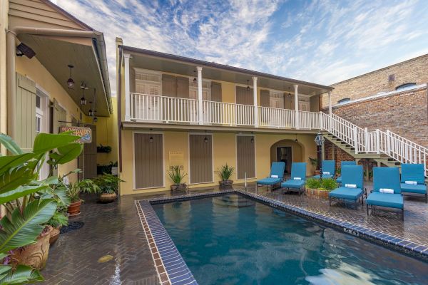 A charming courtyard features a pool, blue lounge chairs, and a two-story building with a balcony under a partly cloudy sky.